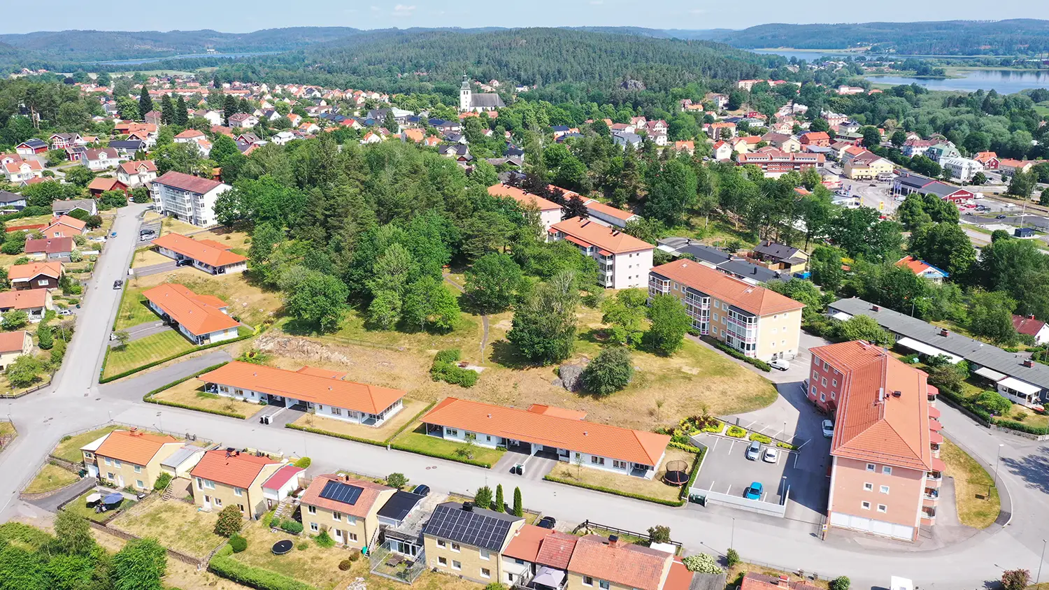 Flygfoto över en liten stad med bostadshus, gröna träd och vägar, omgiven av skogsklädda kullar under en klar himmel.