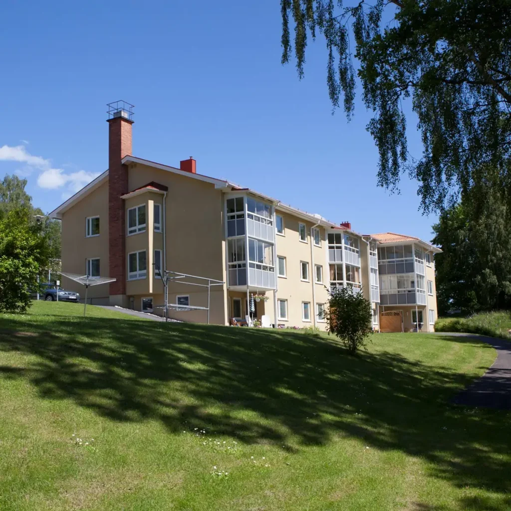 Trevåningshus i beige med balkonger, beläget på en sluttande gräsmatta, omgivet av träd under en klarblå himmel.