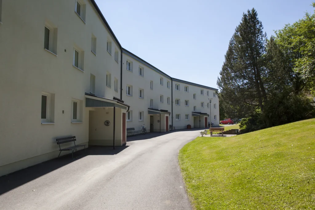 En asfalterad gångväg löper längs ett trevåningshus med vita väggar, bänkar och gräsytor med träd en solig dag.