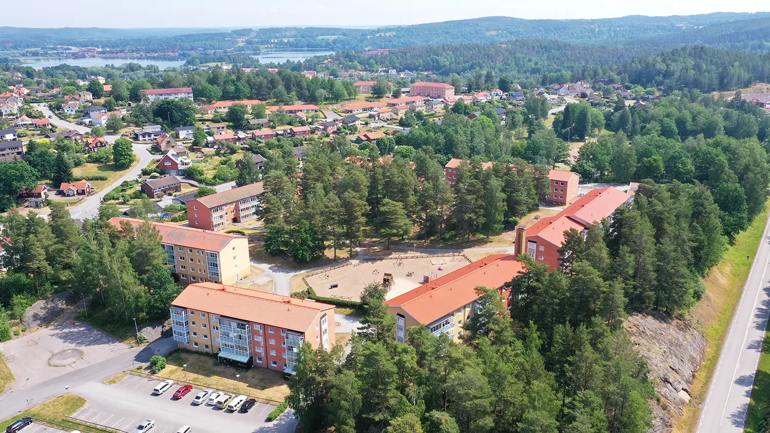 Flygfoto över ett bostadsområde med flerfamiljshus, träd, en lekplats, parkerade bilar och kullar i bakgrunden.