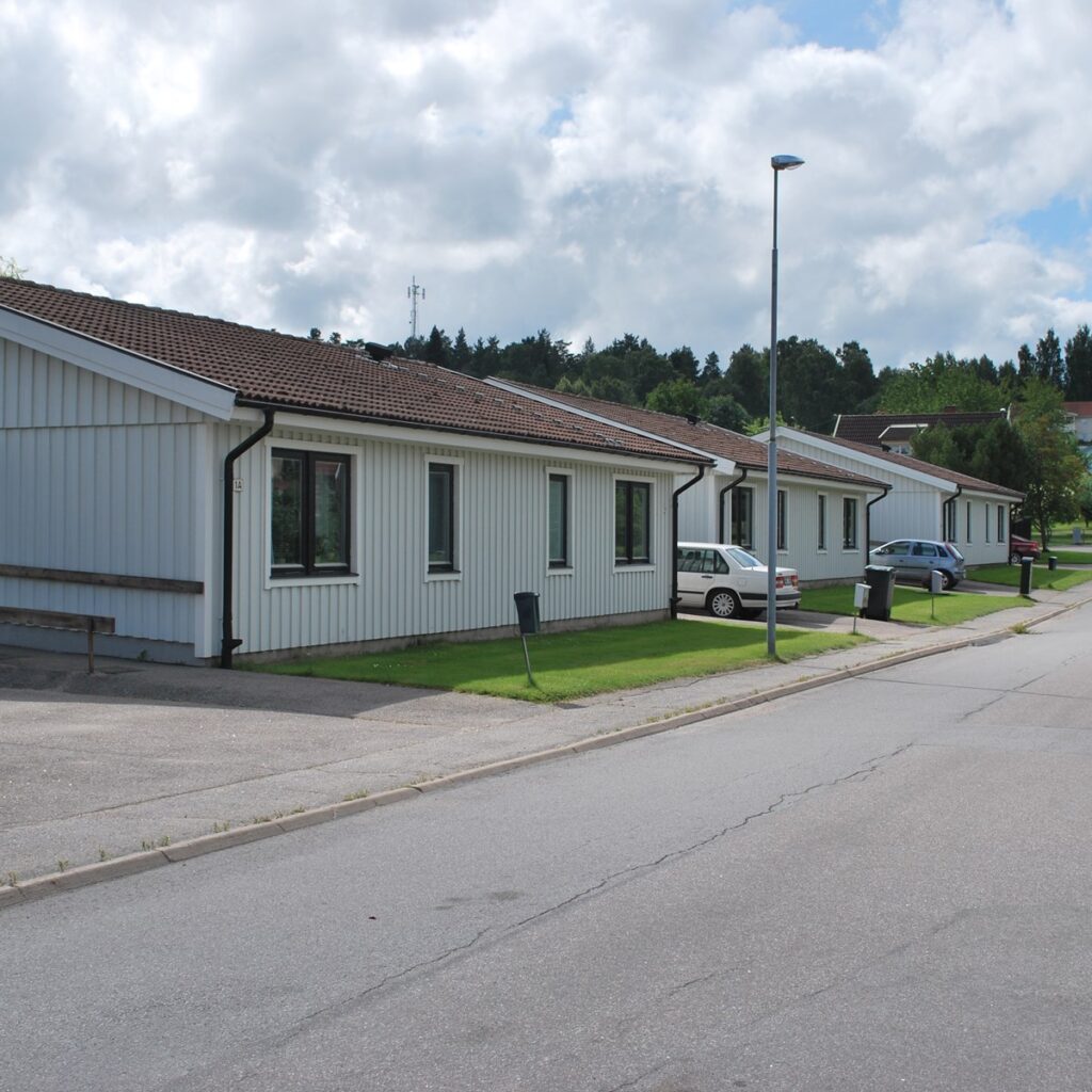 En rad vita envåningshus med bruna tak kantar en lugn bostadsgata med parkerade bilar och gröna gräsmattor under en delvis molnig himmel.