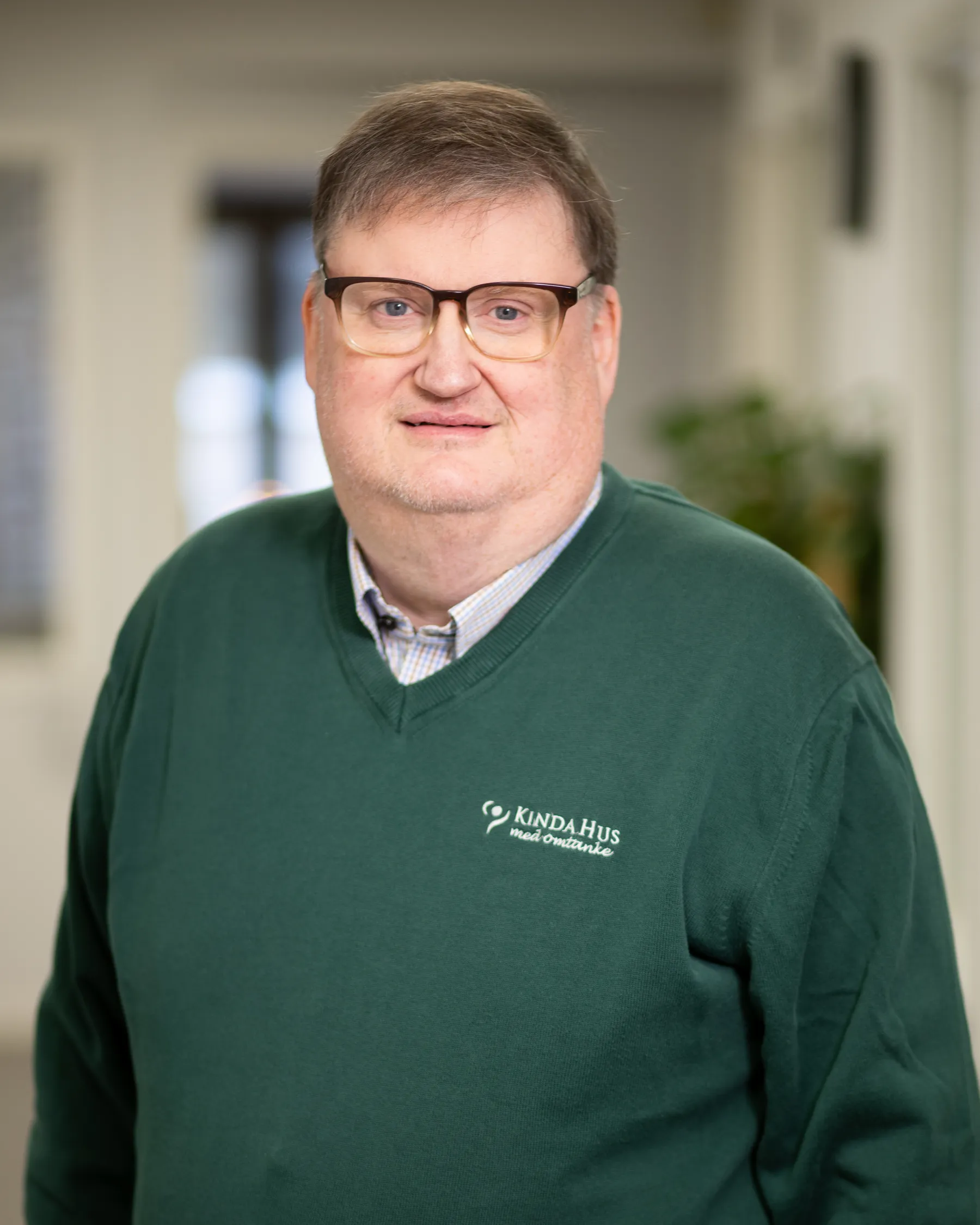 En medelålders man med glasögon och en grön tröja med KINDAHUS-logotypen står inomhus och tittar in i kameran.