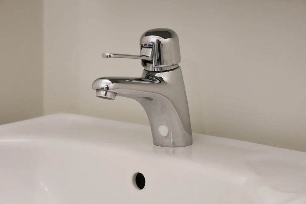 En kromad ettgreppsblandare är monterad på ett vitt handfat med bräddavlopp, mot en vanlig ljus vägg.