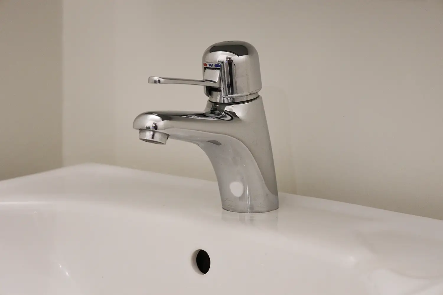 En kromad ettgreppsblandare är monterad på ett vitt handfat med bräddavlopp, mot en vanlig ljus vägg.