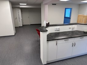 Ett modernt kontorsrum med vita skåp, en svart bänkskiva, ett grått golv och två röda stolar nära en vägg med ordet VÄLKOMMEN.