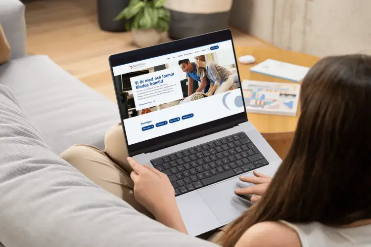 En person sitter i en soffa med en bärbar dator och tittar på en webbplats med svensk text och bilder på människor som arbetar tillsammans.