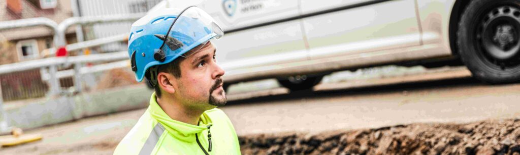 En byggnadsarbetare i varseljacka och blå hjälm tittar upp när han står bredvid ett dike på en arbetsplats, med en vit skåpbil i bakgrunden.