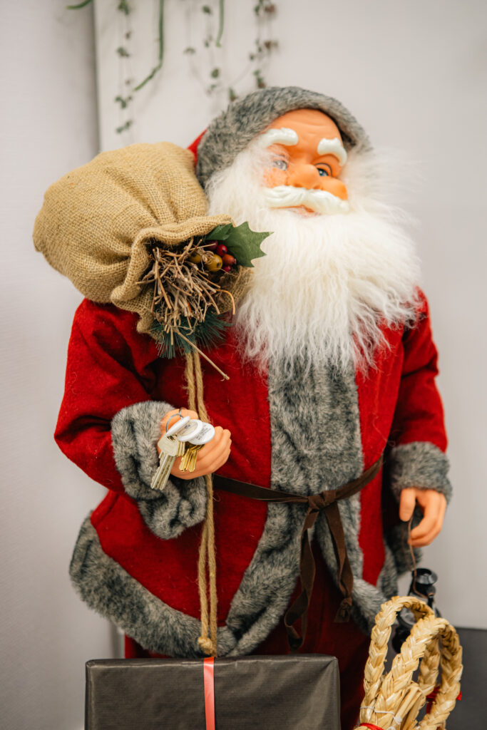 En tomtefigur i röd och grå kostym håller en säck i juteväv och en svart presentförpackning med juldekorationer på säcken.