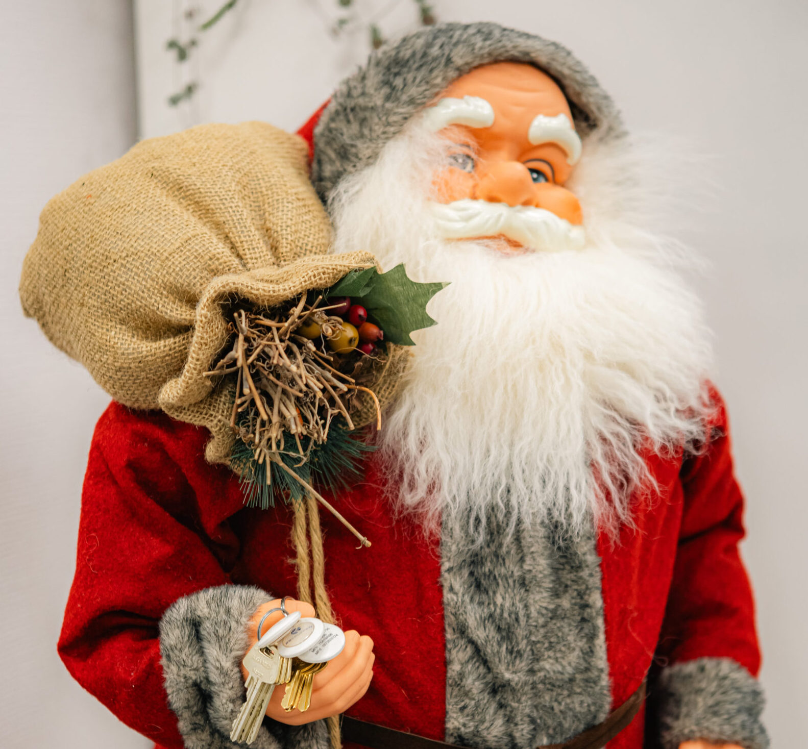 En tomtefigur i röd och grå kostym håller en säck i juteväv och en svart presentförpackning med juldekorationer på säcken.