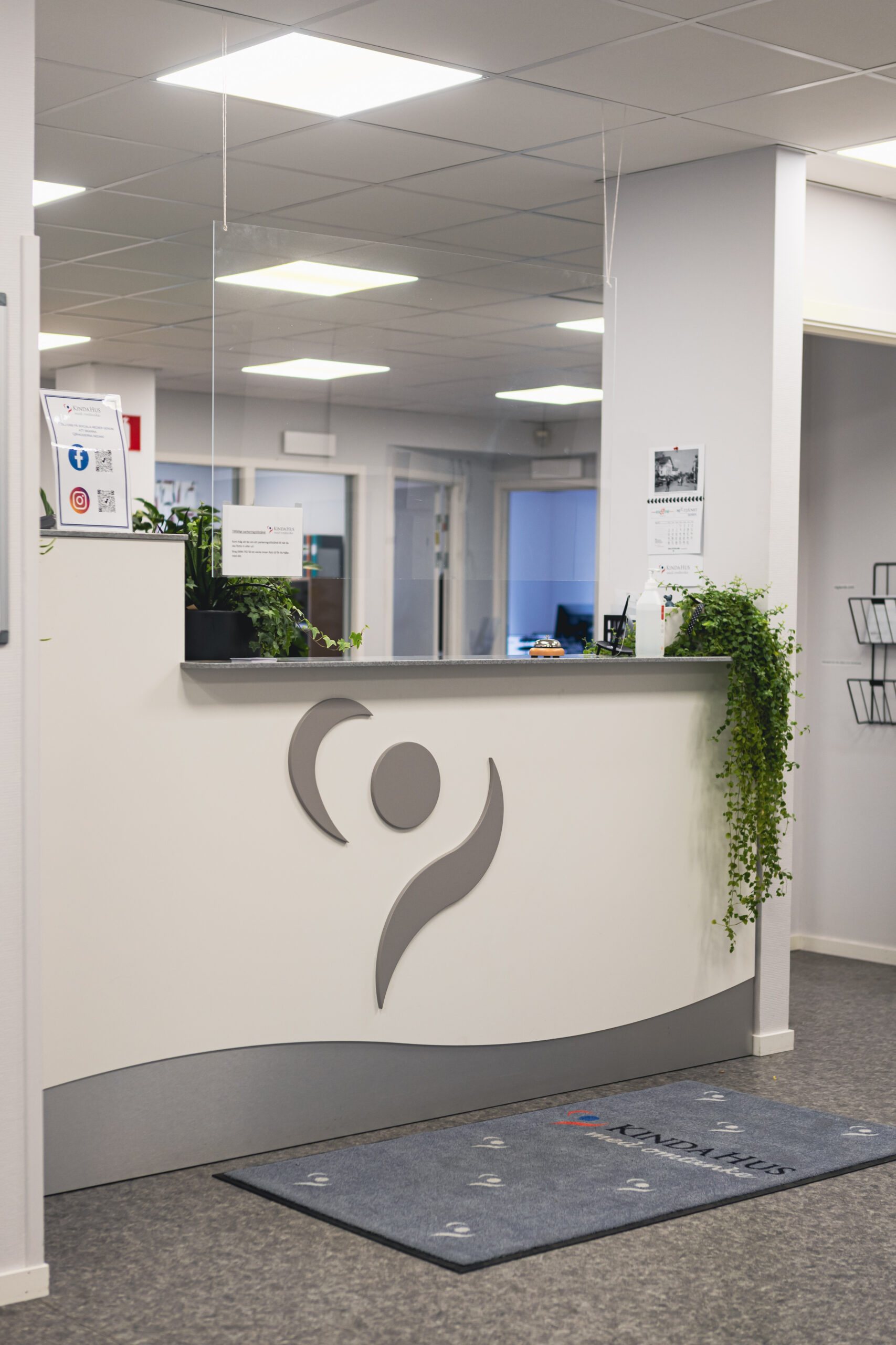 En modern receptionsdisk med en minimalistisk logotyp, krukväxter och informationsblad i ett väl upplyst kontorsutrymme.