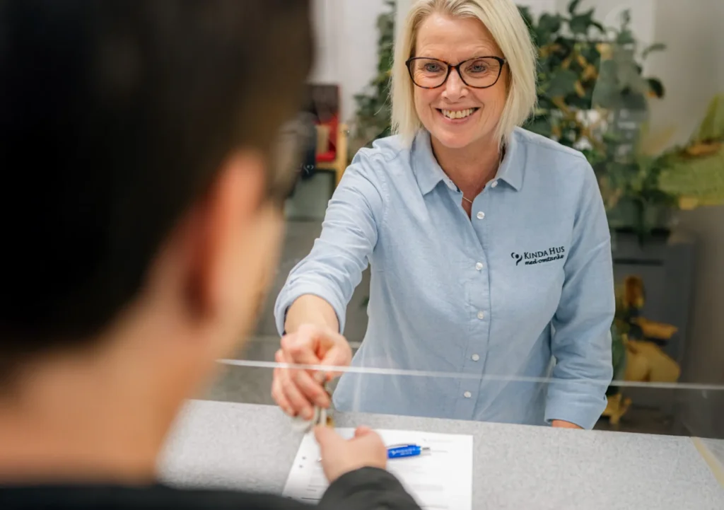 En kvinna med glasögon och blå skjorta ler när hon överlämnar ett dokument till en person på andra sidan en disk med pappersarbete och penna synliga.