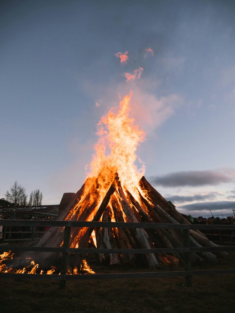 En stor brasa gjord av staplade stockar brinner starkt i ett inhägnat utomhusområde i skymningen, med en folkmassa i bakgrunden.
