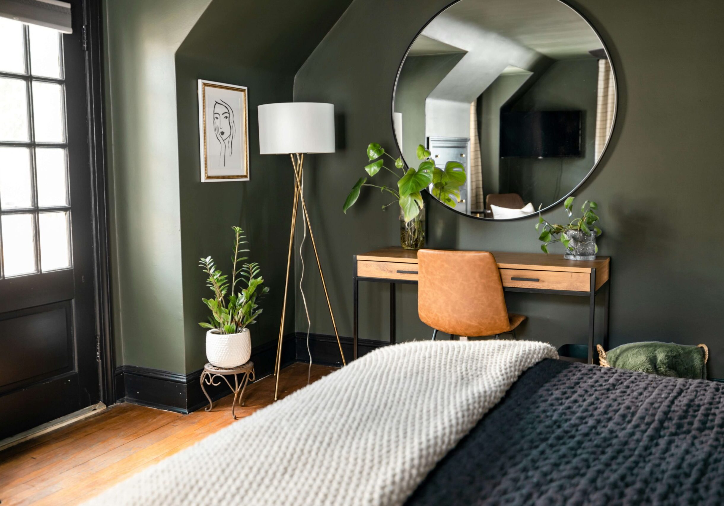 Ett modernt sovrum med mörkgröna väggar, en stor rund spegel ovanför ett träskrivbord, en brun stol, krukväxter och mysiga texturerade filtar - ett stilfullt utrymme som varje hyresvärd i Kinda skulle vilja erbjuda.